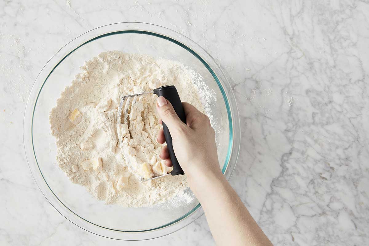 Hand using a pastry cutter to cut butter into a bowl of flour 