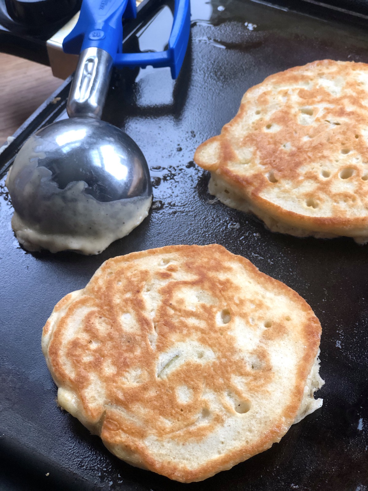Two pancakes cooking on a griddle, muffin scoop on the side.