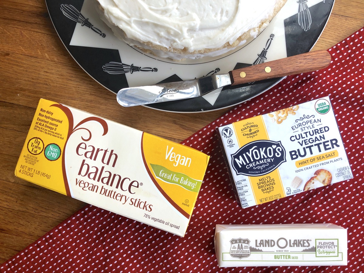 Containers of Earth Balance, Miyoko's, and Lane O Lakes butters on a wooden table, frosted valilla cake alongside.