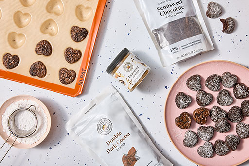 Heart shaped brownies in a sil pat mold with recipe ingredients and baked brownies on a pink plate.
