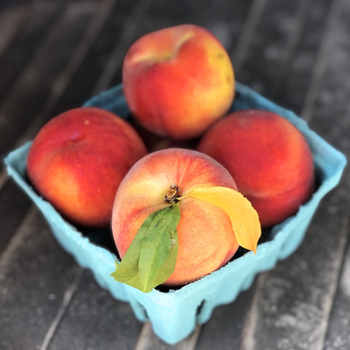 A quart container of beautiful fresh peaches, one with the leaves intact.