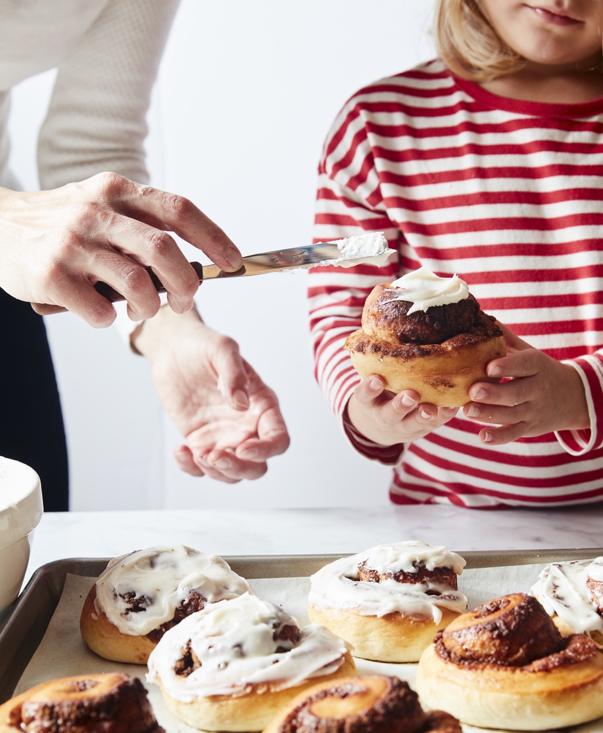 Little girl in a red-striped shirt holding a cinnamon roll while Mom frosts it with white icing.