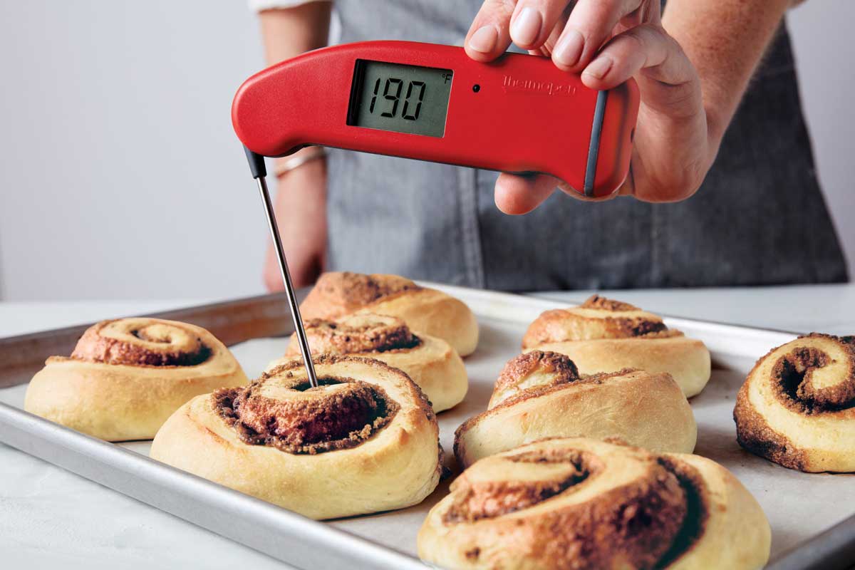 Baked cinnamon rolls on a baking sheet, one's temperature being taken with a probe thermometer and showing 190°F.