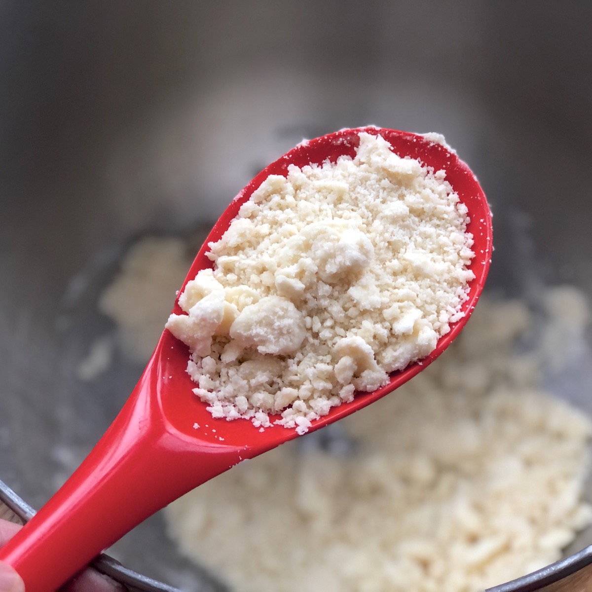 Butter and bacon fat worked into a bowl of dry ingredients to create a crumbly mixture, shown pictured in the bowl of a red spoon.