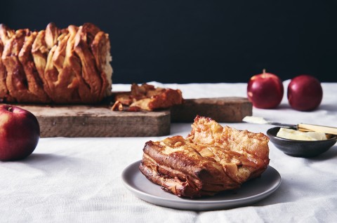 A loaf of Cinnamon Apple Pull-Apart Bread next to a few apples and slice of pull-apart bread on a plate, ready to be eaten. - select to zoom