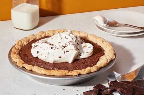 A Chocolate Cream Pie topped with whipped cream next to a stack of plates and a glass of milk - select to zoom
