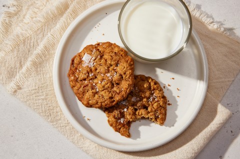 An overhead photo of two Oatmeal Cookies on small plate along with a glass of milk. The cookies have crackly tops and flaky sea salt sprinkled on them, one cookie has a bite taken out of it.  - select to zoom