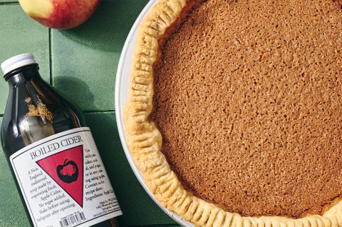 An apple pie seen from above with a smooth texture and a bottle of boiled cider laying on the table next to it. - select to zoom