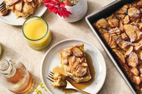 A slice of Apple French Toast Bake on a light colored plate with syrup spilling out beneath the french toast. The scene also included a cup of orange juice, syrup in a jar, and shows the corner of the baked french toast in a baking pan. - select to zoom