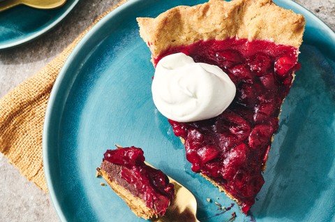 Cranberry-Fudge Pie seen from above with dark and glossy cranberries, a dollop of whipped cream, and a beautifully browned crust. A bite on a fork show the fudgy bottom of the pie. - select to zoom
