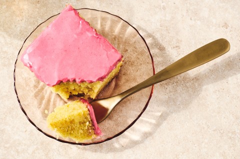 A slice of Cranberry-Orange Olive Oil Cake on a glass plate with a bite on a fork showing a moist golden cake and pastel pink frosting. - select to zoom