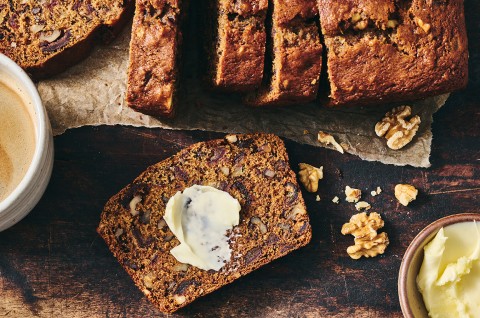 A slice of buttered Old-Fashioned Date Nut Bread on a plate atop a dark counter and with a latte and sliced loaf in the corner of the photo. - select to zoom