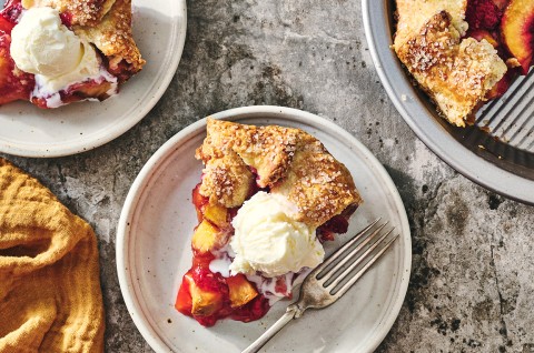 A slice of Rustic Raspberry-Peach Pie topped with vanilla ice cream on a white plate with a fork next to it. - select to zoom