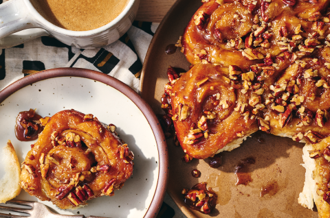 A single sticky bun on a plate adjacent to freshly baked buns. - select to zoom
