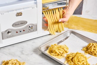 Cutting pasta into noodles with an electric pasta machine, next to a sheet pan of fresh pasta noodles