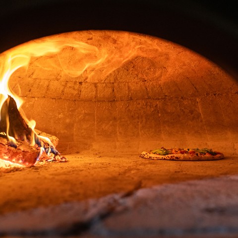 Interior view of a wood fired pizza oven with a pizza baking in the corner and a wood fire on the other side. - select to zoom