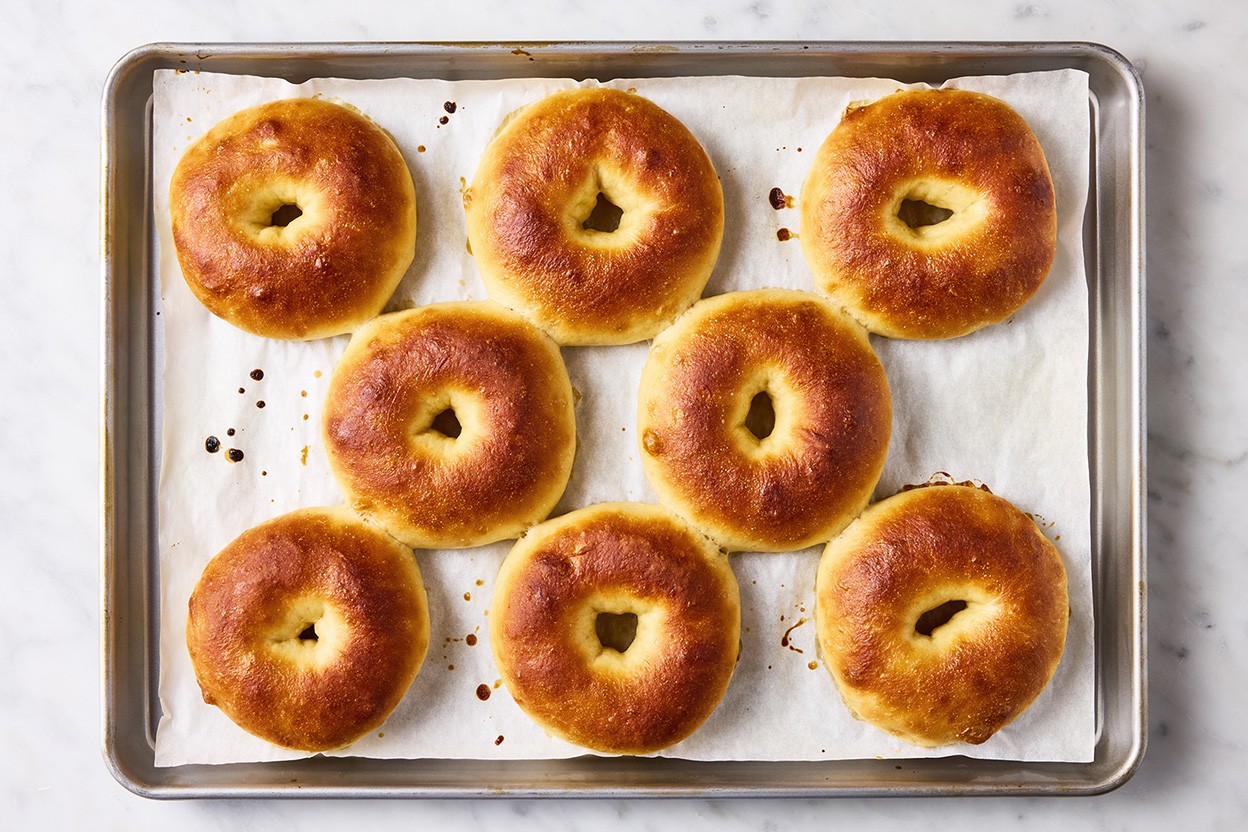 Eight beautifully baked classic bagels on a sheet tray and parchment paper.