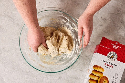 Hands mixing bagel dough in a glass bowl.