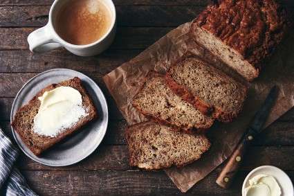 A loaf of Sourdough Banana Bread cut into slices, with one piece slathered in butter