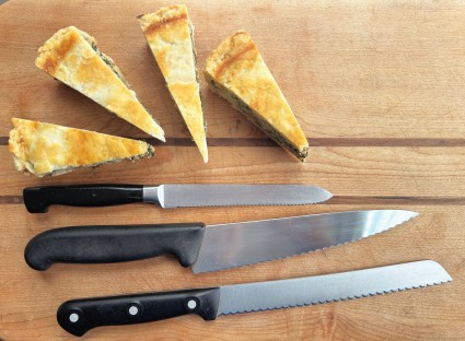 Three sizes of serrated knife on a cutting board, accompanied by several slices of a flaky vegetable tart.