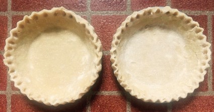Two pie crusts with tall crimped edges, side by side, ready to go into the oven for prebaking.