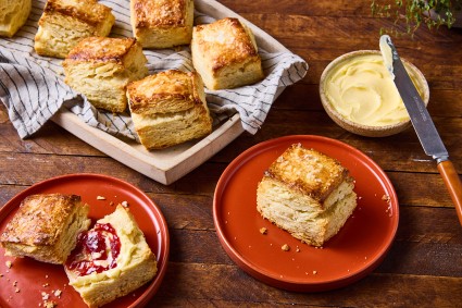 A tray of tall flaky biscuits, a plate with a single rectangular biscuit, a bowl of butter, and another red plate with a sliced biscuit that has jelly and butter on it.