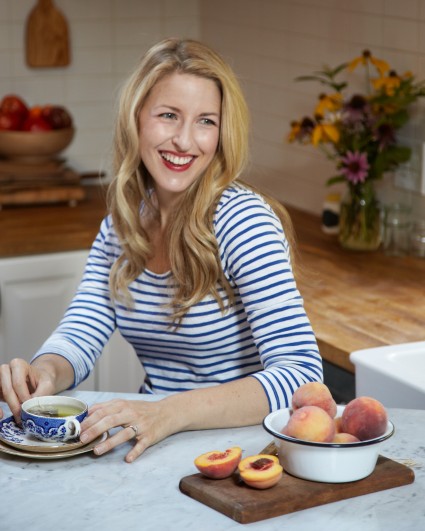 A photo of Sarah Copeland in a kitchen drinking coffee next to a bowl of peaches