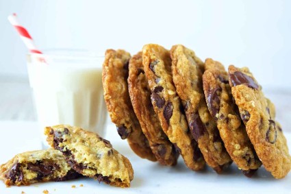 A side angle photo of oatmeal chocolate chip cookies leaning against a glass of milk. The cookies have crisp edges and chewy centers with lots of chocolate chips. 