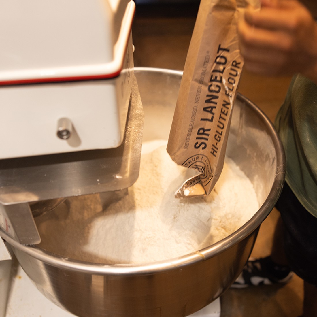 Dough being made in a stand mixer with a person pouring in King Arthur Baking's Lancelot Flour. - select to zoom