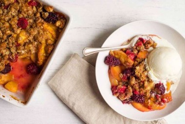 A pan of freshly baked, crumb-topped Summer Fruit Crisp next to a bowl of fruit crisp with a spoon
