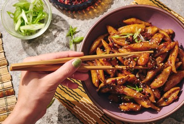A person picking up hand cut noodles dressed with a dark umami sauce using chopsticks.