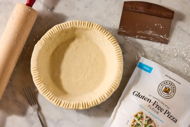 A rolling pin, pie crust, pastry knife, and bag of King Arthur Gluten-Free Pizza Flour shown on a counter and neatly arranged.