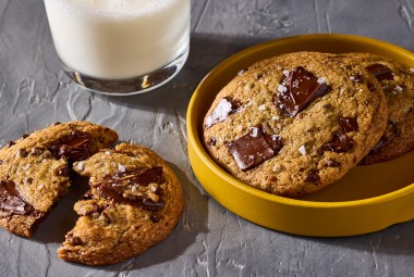 Beautifully baked High-Altitude Chocolate Chip Cookies shown on a plate and counter with a glass of milk in the background.