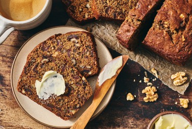 A slice of buttered Old-Fashioned Date Nut Bread on a plate atop a dark counter and with a latte and sliced loaf in the corner of the photo.