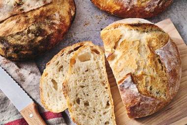 Fresh baked loaves of No-Knead Crusty White Bread on a table with jam and butter and slices on a cutting board.