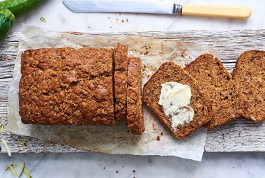 An overhead photo of a loaf of zucchini bread. A few slices have been cut off the end, showing the tender interior crumb. One slice has softened butter spread on it. 